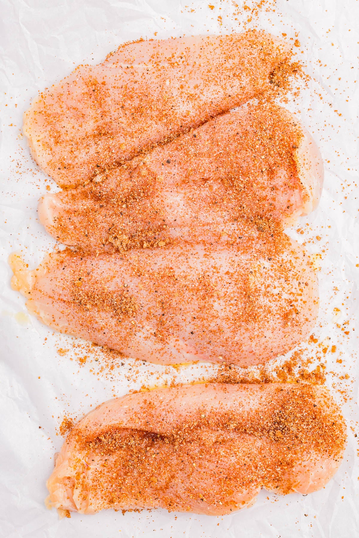 Four raw chicken breasts laid flat on white parchment paper, evenly sprinkled with a reddish-brown spice rub.