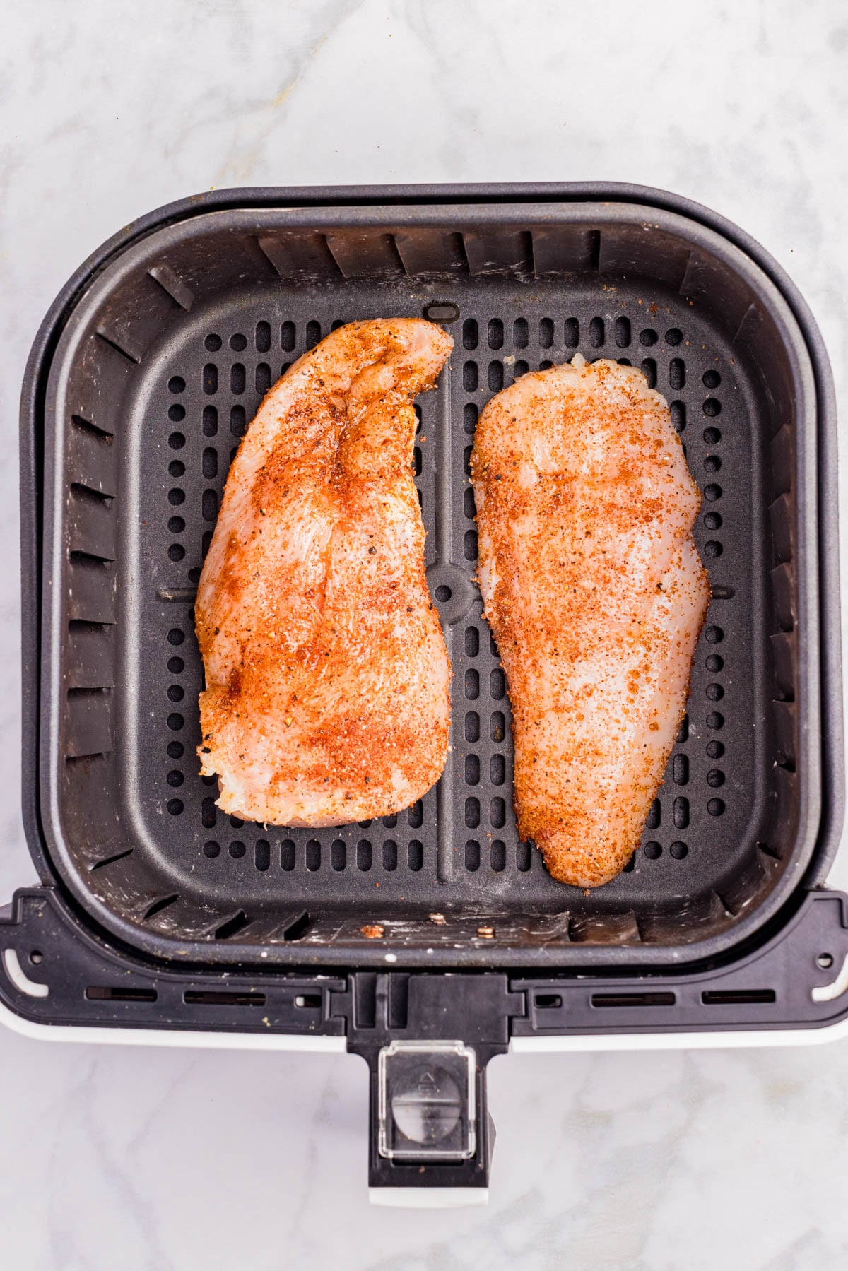 Two raw, seasoned chicken breasts placed side by side in the basket of an air fryer on a marble countertop.
