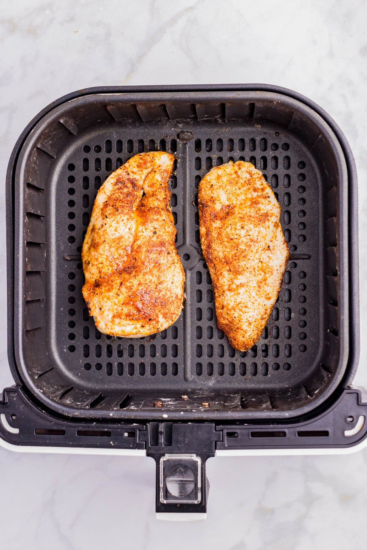 Two seasoned chicken breasts placed side by side in the basket of an air fryer on a marble surface.