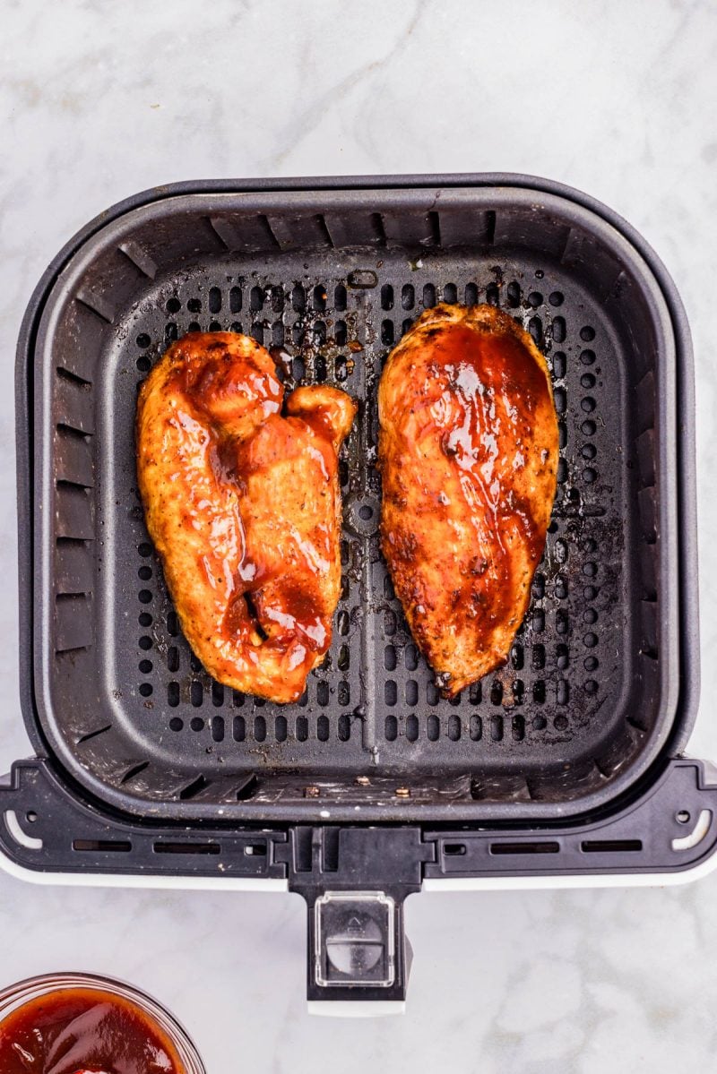 Two pieces of cooked, sauced chicken breast in an air fryer basket, photographed from above on a light countertop.