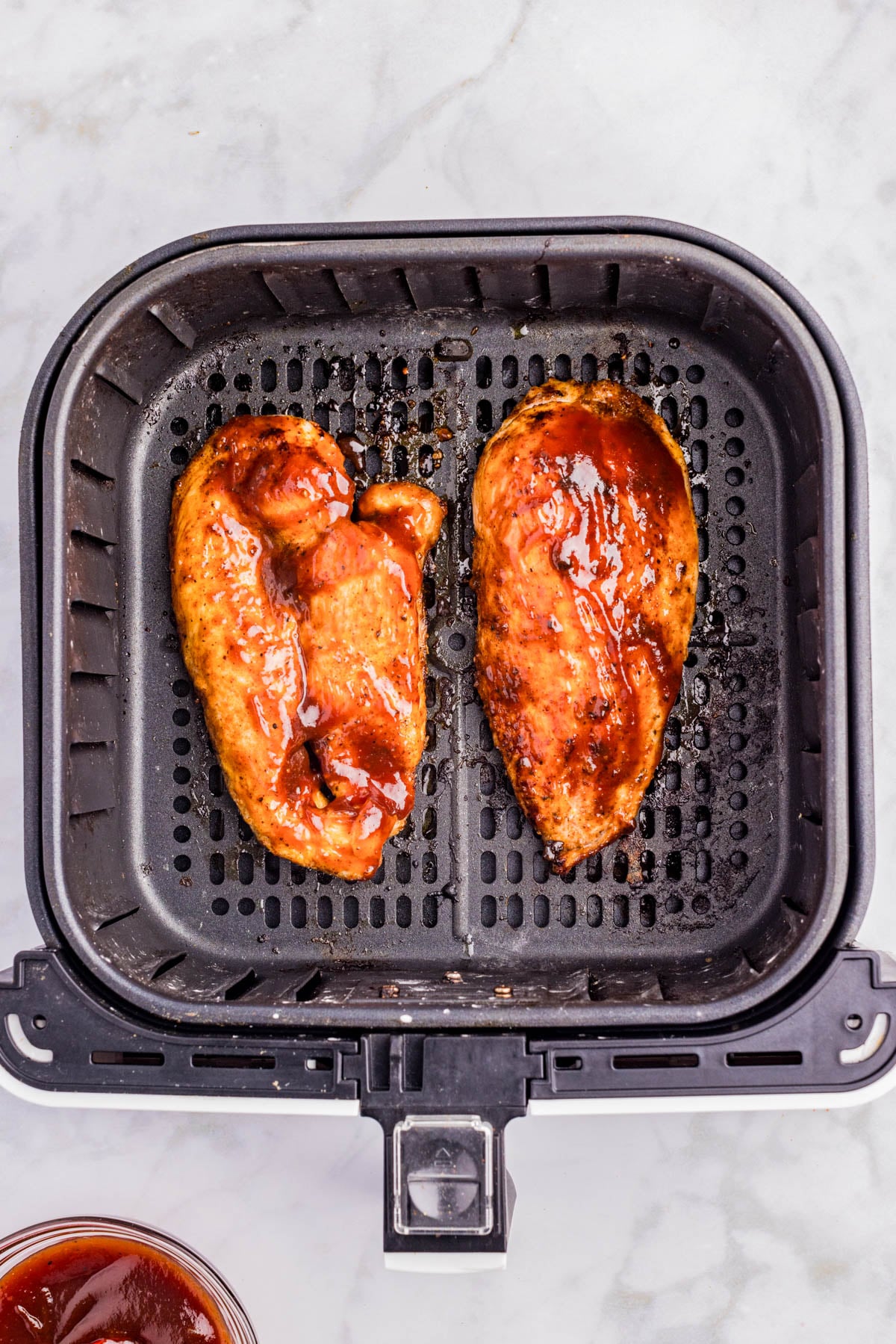 Two pieces of cooked, sauced chicken breast in an air fryer basket, photographed from above on a light countertop.