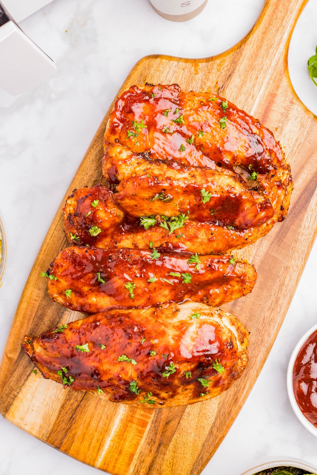 Four grilled chicken breasts coated in barbecue sauce and garnished with chopped parsley, arranged on a wooden serving board.