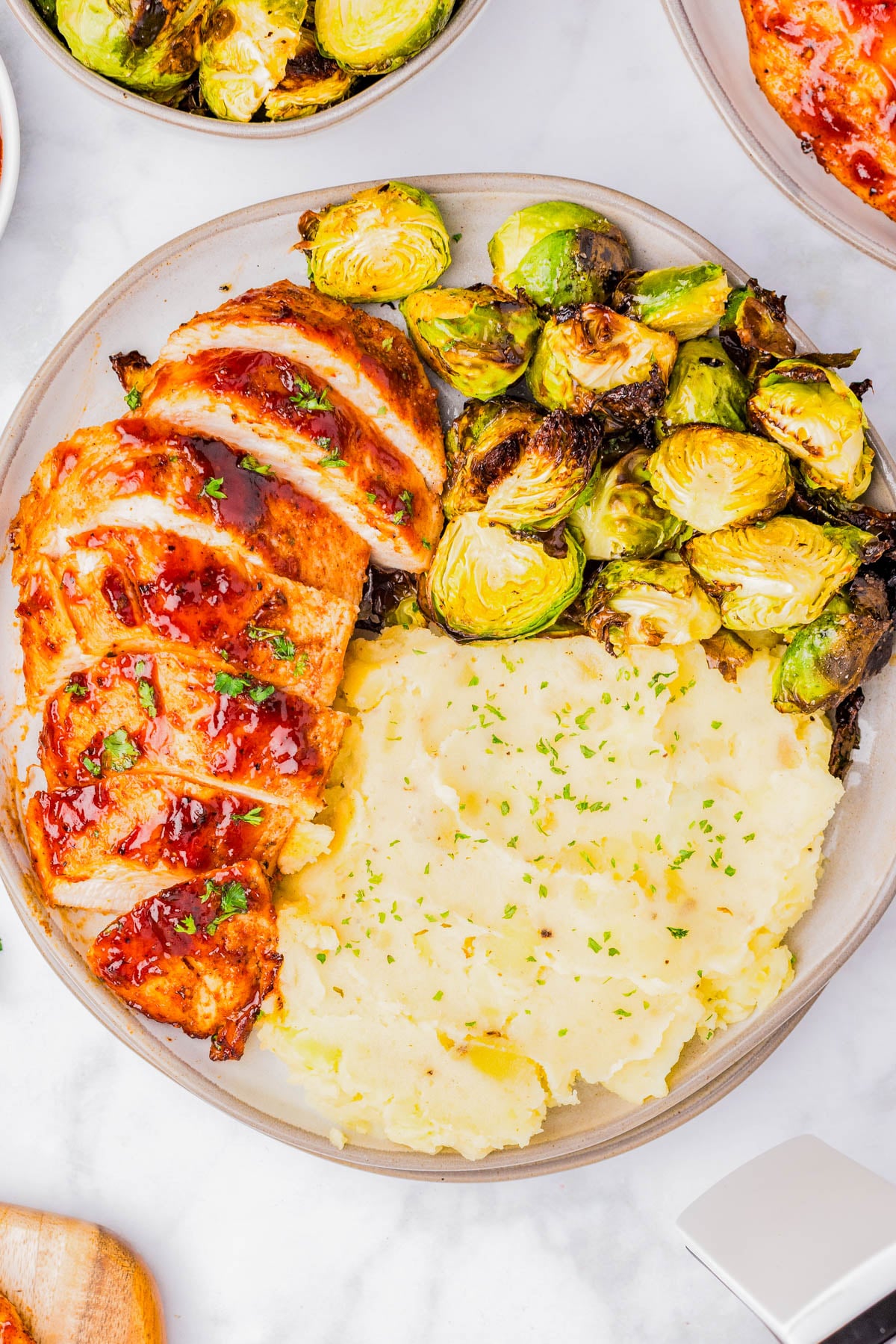 A plate with sliced glazed chicken breast, roasted Brussels sprouts, and a serving of mashed potatoes garnished with herbs.