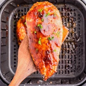 A wooden spatula holds a glazed, cooked chicken breast over an air fryer basket.