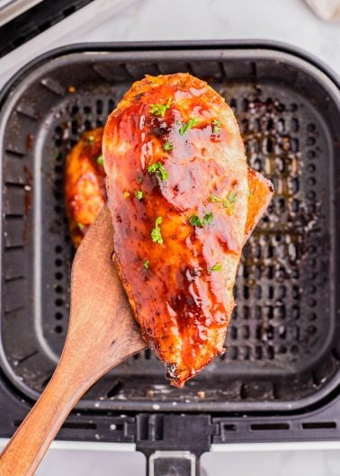 A wooden spatula holds a glazed, cooked chicken breast over an air fryer basket.