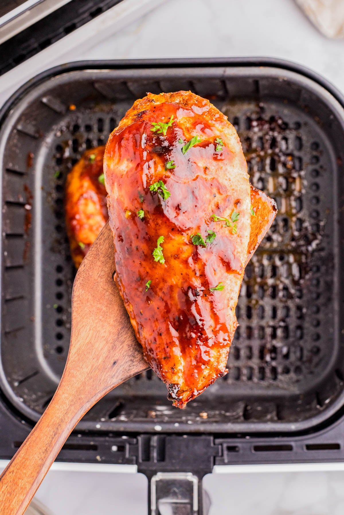 A wooden spatula holds a glazed, cooked chicken breast over an air fryer basket.
