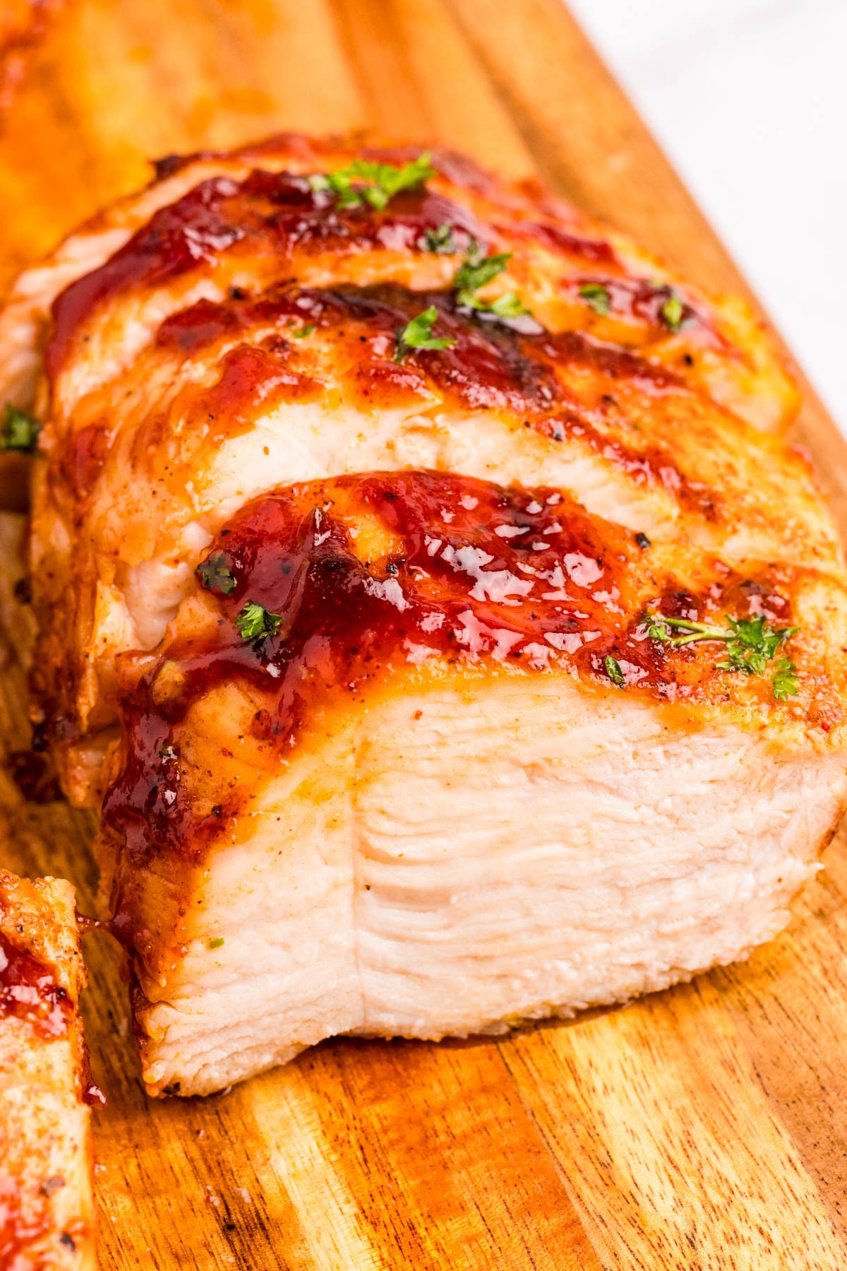 Sliced cooked chicken breast with a glazed sauce and chopped herbs on a wooden cutting board.