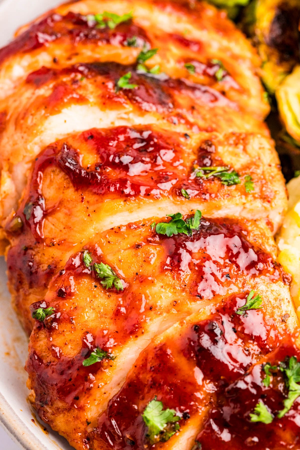 Sliced glazed chicken breast garnished with chopped parsley, served next to vegetables on a white plate.