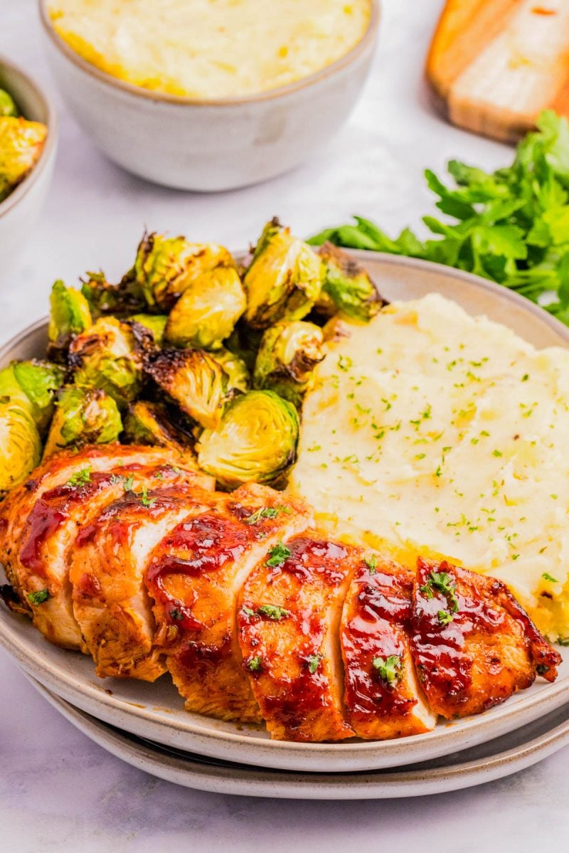 A plate with sliced glazed chicken breast, roasted Brussels sprouts, and mashed potatoes garnished with herbs.