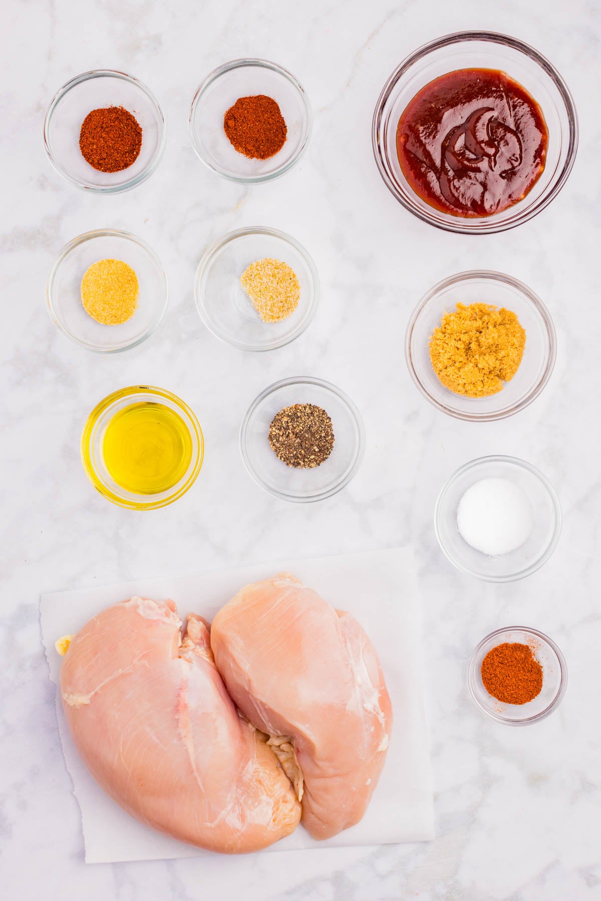 Two raw chicken breasts on parchment surrounded by small glass bowls containing oil, spices, brown sugar, salt, pepper, and barbecue sauce on a white surface.
