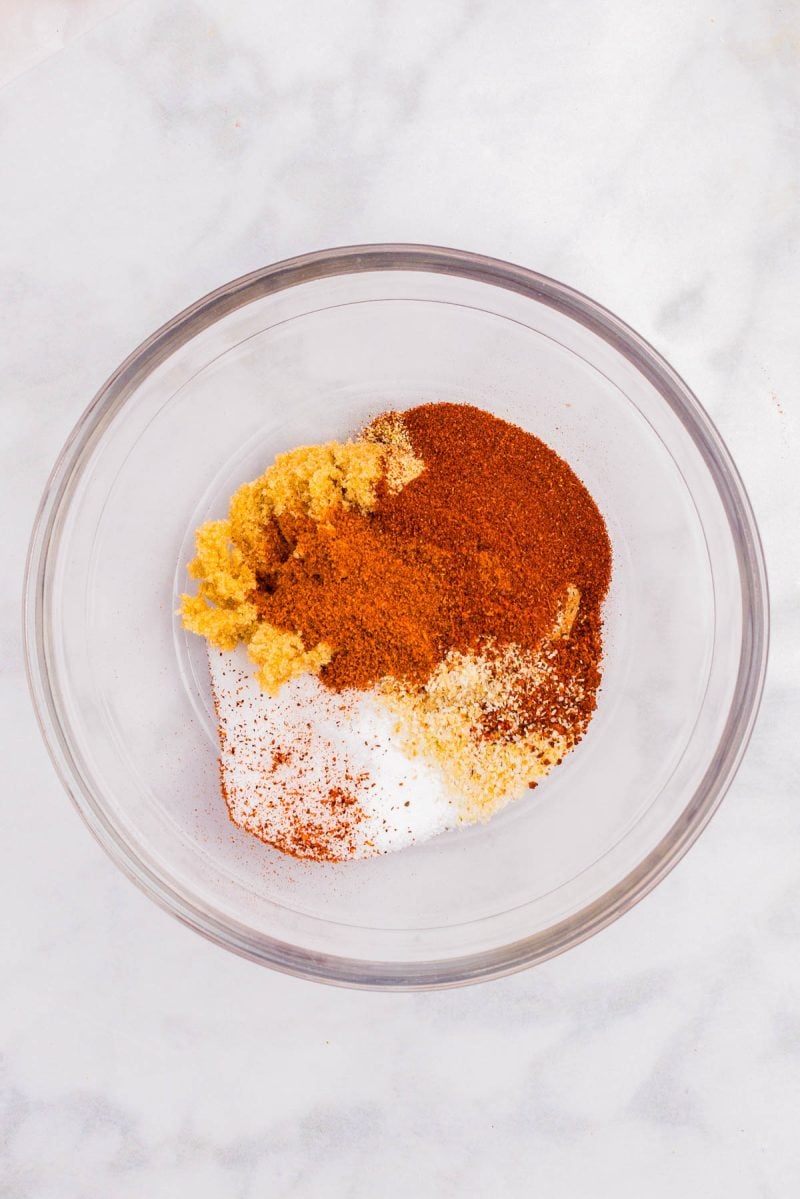 A glass bowl containing separate piles of various ground spices and brown sugar on a white surface.