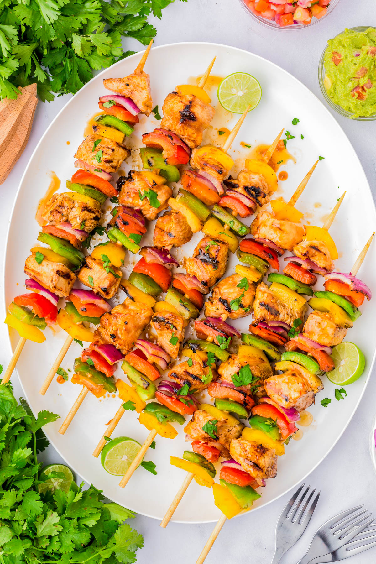 Grilled chicken and vegetable skewers arranged on a white platter, garnished with chopped herbs and lime wedges, surrounded by fresh cilantro and dipping sauces.