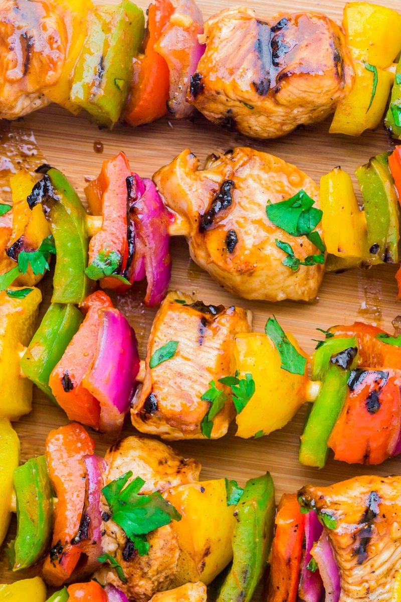 Close-up of grilled kebabs with pieces of chicken, red and green bell peppers, yellow peppers, and red onions garnished with chopped parsley on a wooden surface.