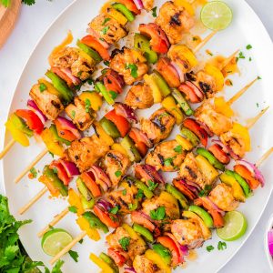Grilled chicken and vegetable skewers arranged on a white platter, garnished with lime slices and fresh herbs, with cilantro and dips on the side.