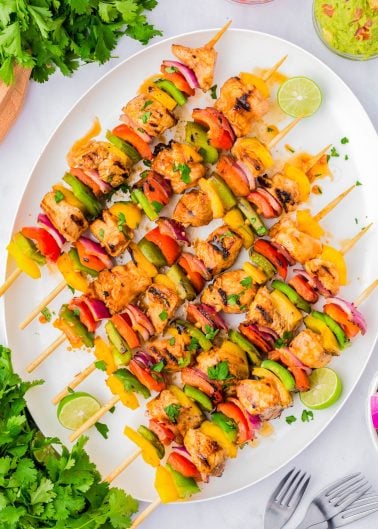 Grilled chicken and vegetable skewers arranged on a white platter, garnished with lime slices and fresh herbs, with cilantro and dips on the side.
