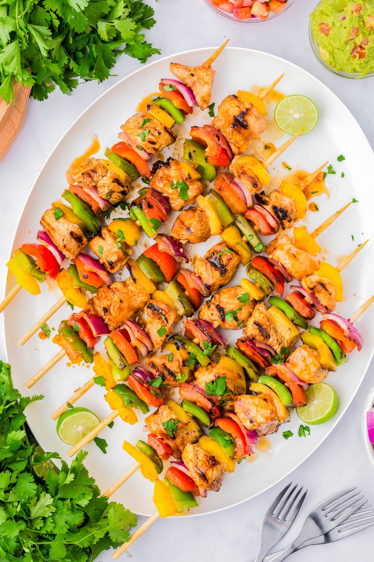 Grilled chicken and vegetable skewers arranged on a white platter, garnished with lime slices and fresh herbs, with cilantro and dips on the side.