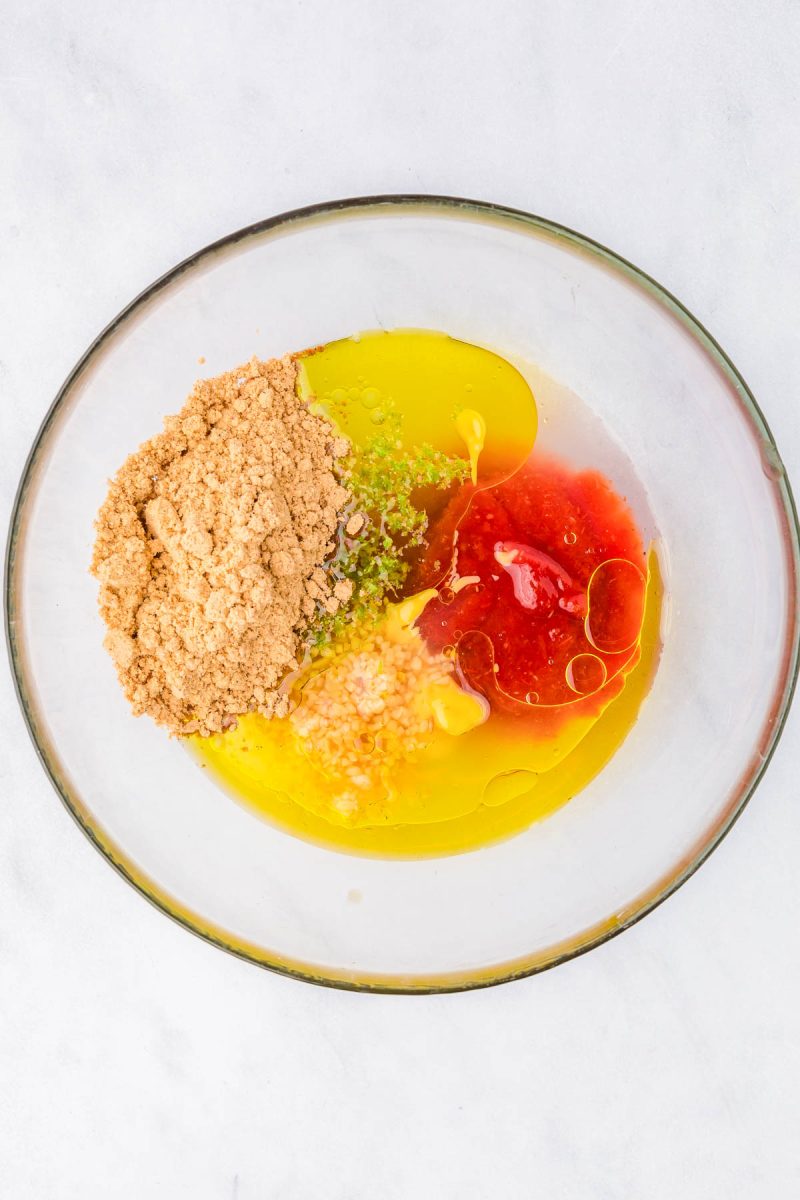 A glass bowl containing olive oil, brown sugar, minced garlic, lime zest, and red sauce on a white surface.