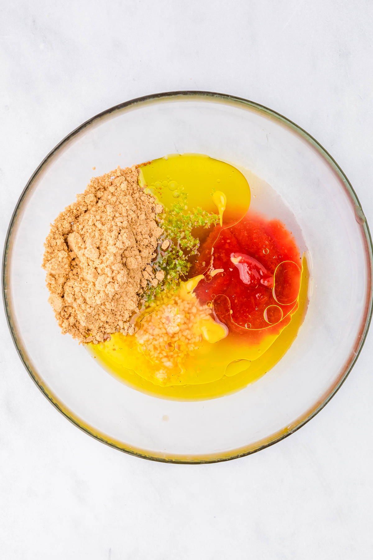 A glass bowl containing olive oil, brown sugar, minced garlic, lime zest, and red sauce on a white surface.
