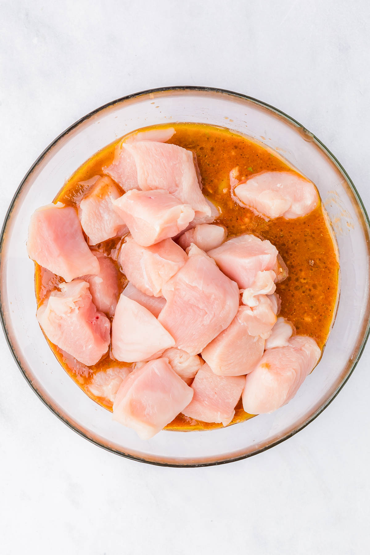 Raw chicken pieces in a glass bowl with orange marinade, viewed from above on a white surface.
