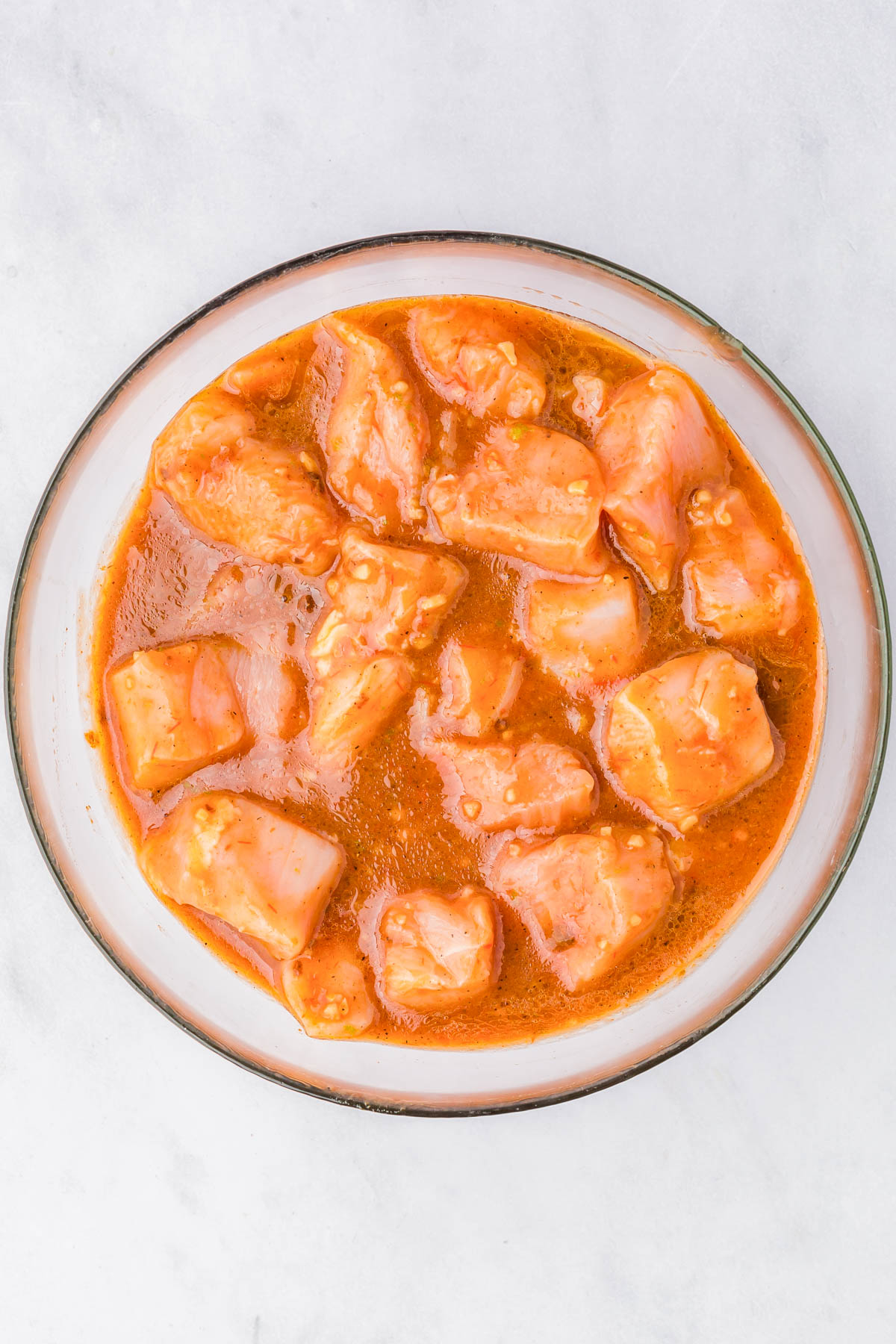 A bowl containing raw chicken pieces marinating in an orange-colored sauce on a white surface.