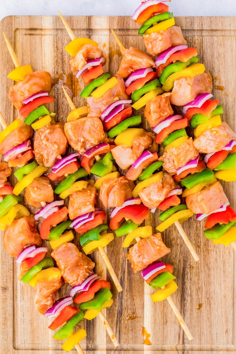 Wooden skewers with marinated chicken, green and yellow bell peppers, and red onion pieces, arranged on a wooden cutting board.