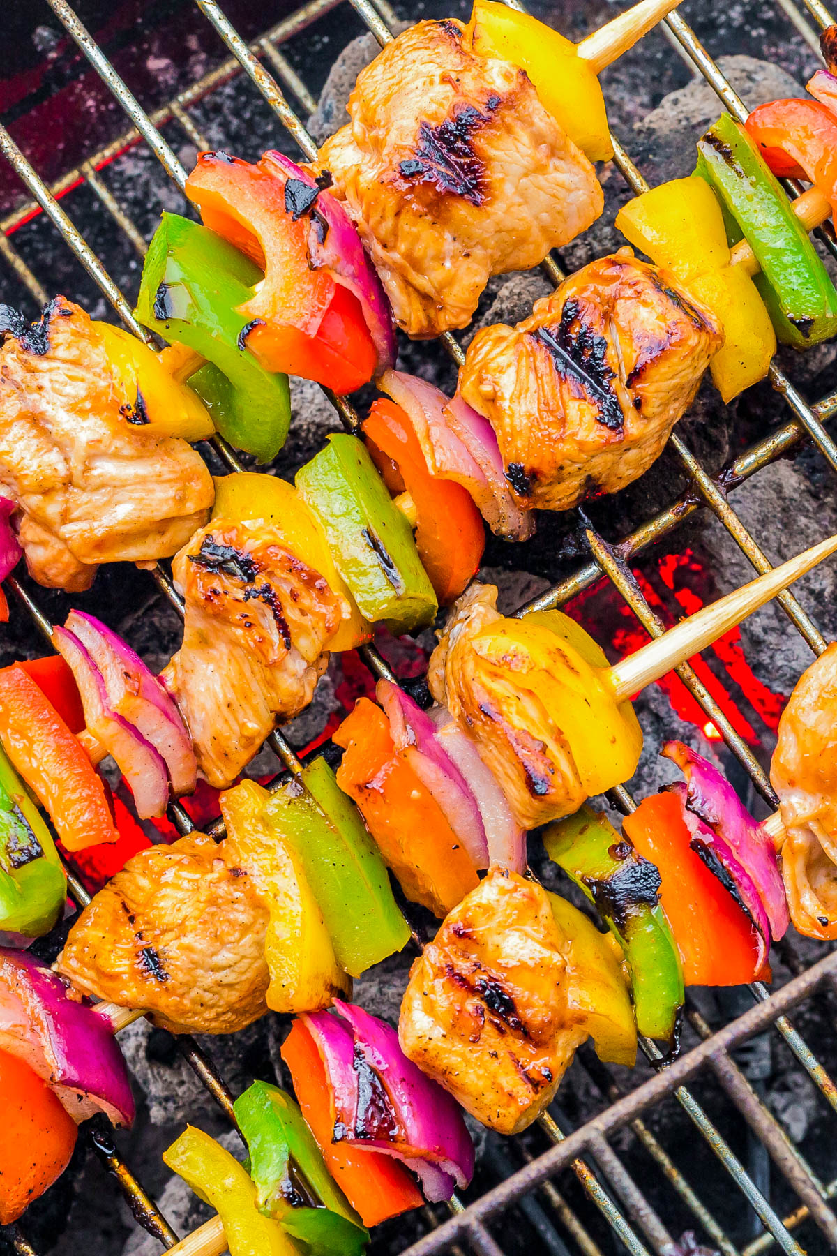 Grilled skewers with pieces of chicken, red onion, yellow bell pepper, green bell pepper, and red bell pepper on a barbecue grill over hot coals.