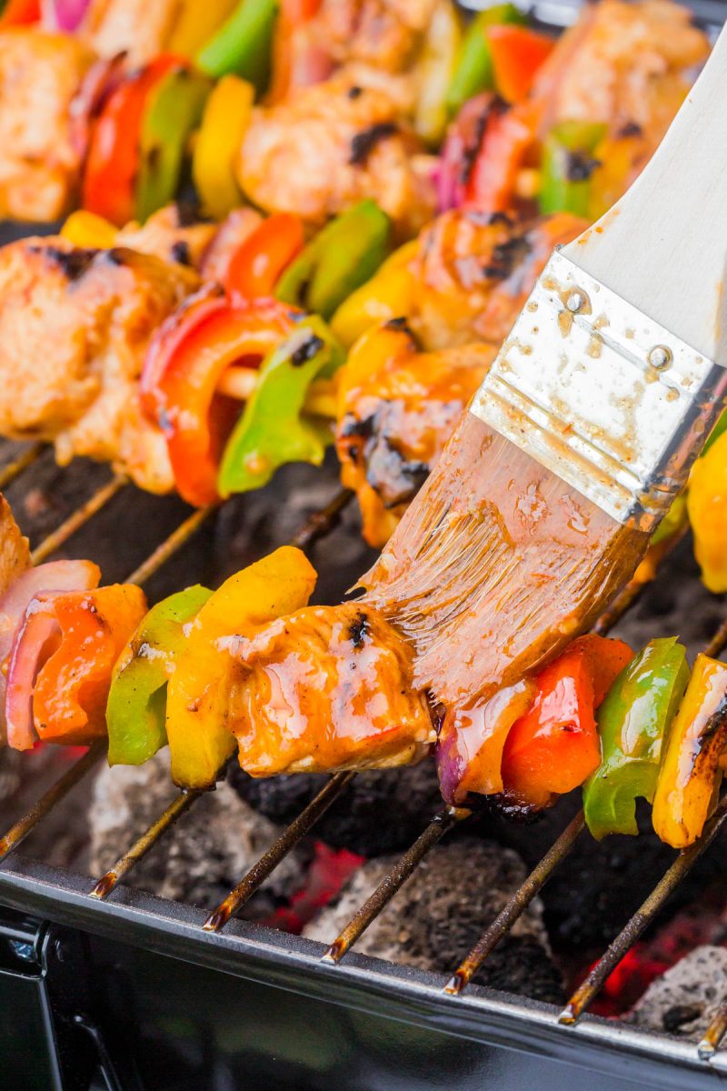 A close-up of vegetable and meat skewers on a grill, with a brush applying sauce to the kebabs.