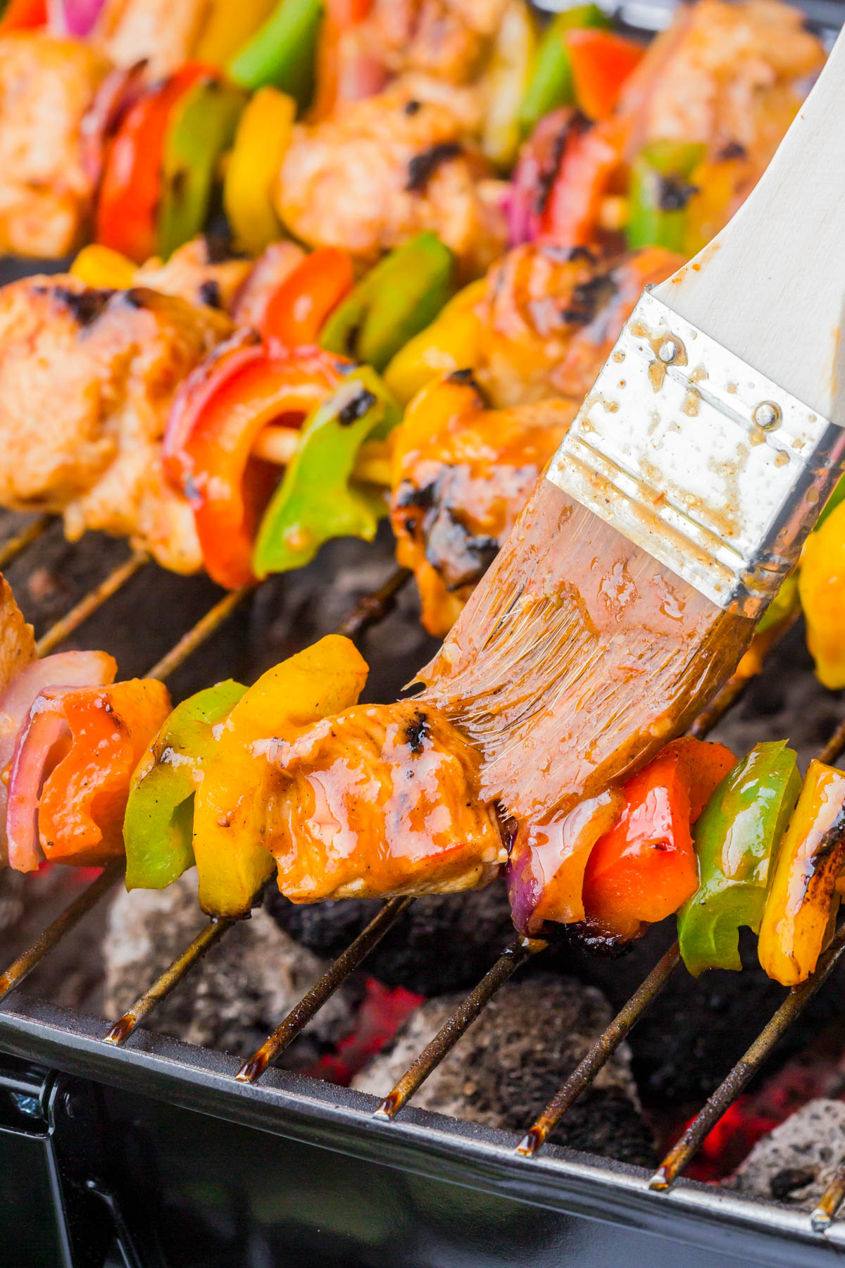 A close-up of vegetable and meat skewers on a grill, with a brush applying sauce to the kebabs.