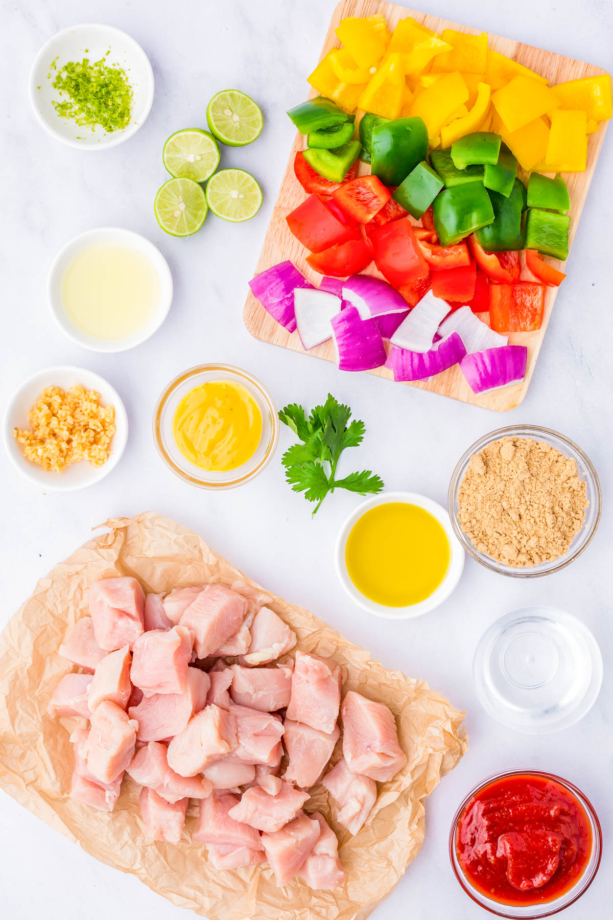Ingredients for a chicken recipe arranged on a white surface, including raw diced chicken, chopped bell peppers, red onion, sauces, lime, garlic, and seasonings.