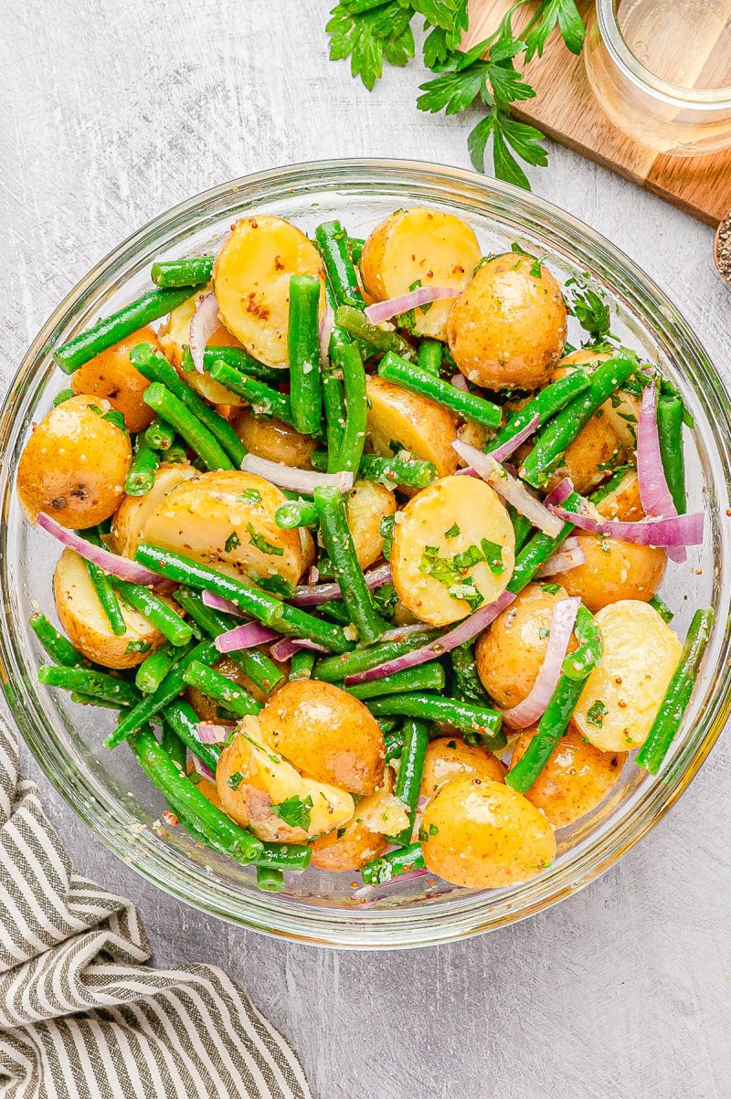 A glass bowl filled with potato salad featuring baby potatoes, green beans, red onion slices, and chopped herbs on a light surface.
