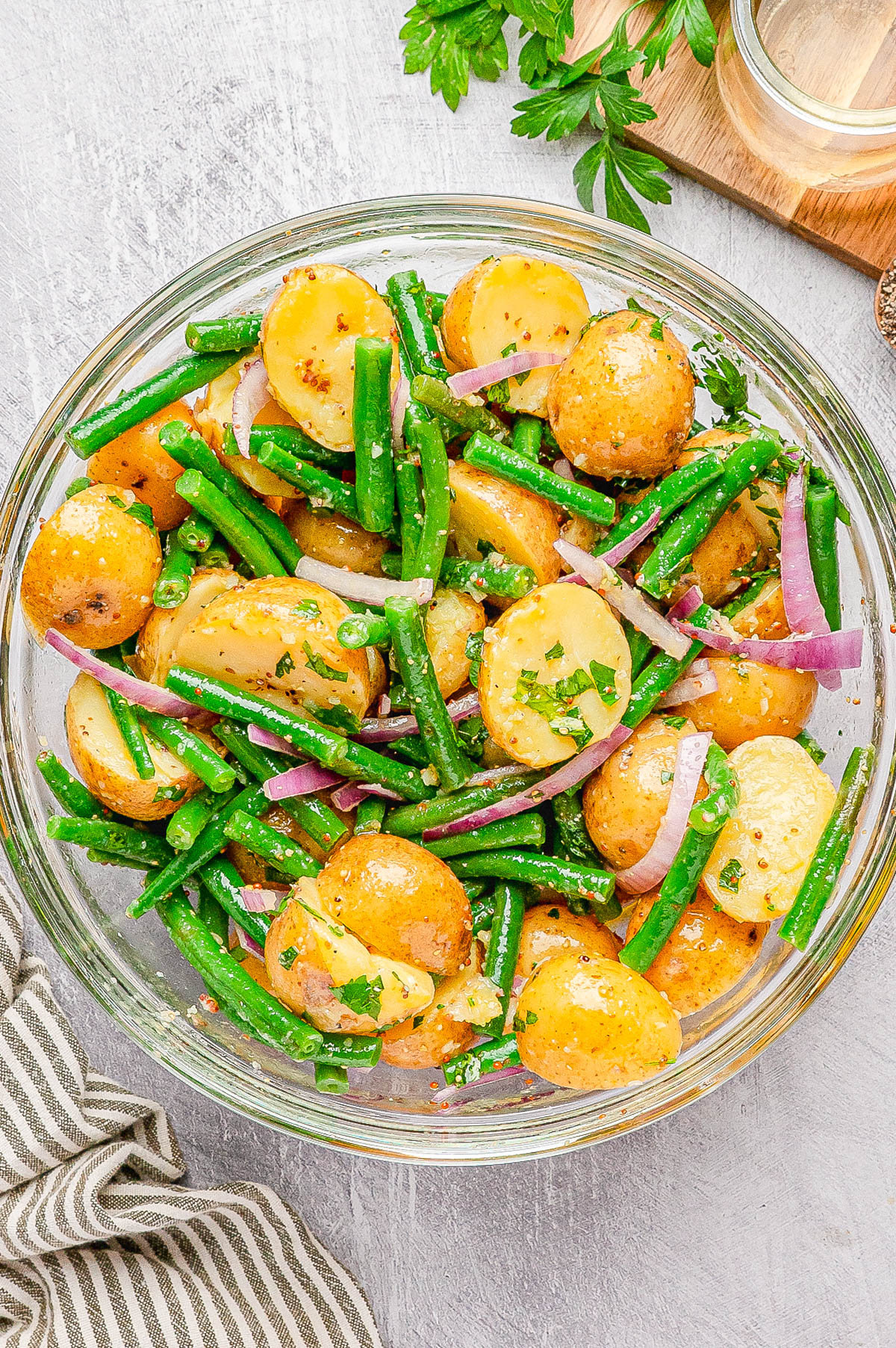 A glass bowl filled with potato salad featuring baby potatoes, green beans, red onion slices, and chopped herbs on a light surface.