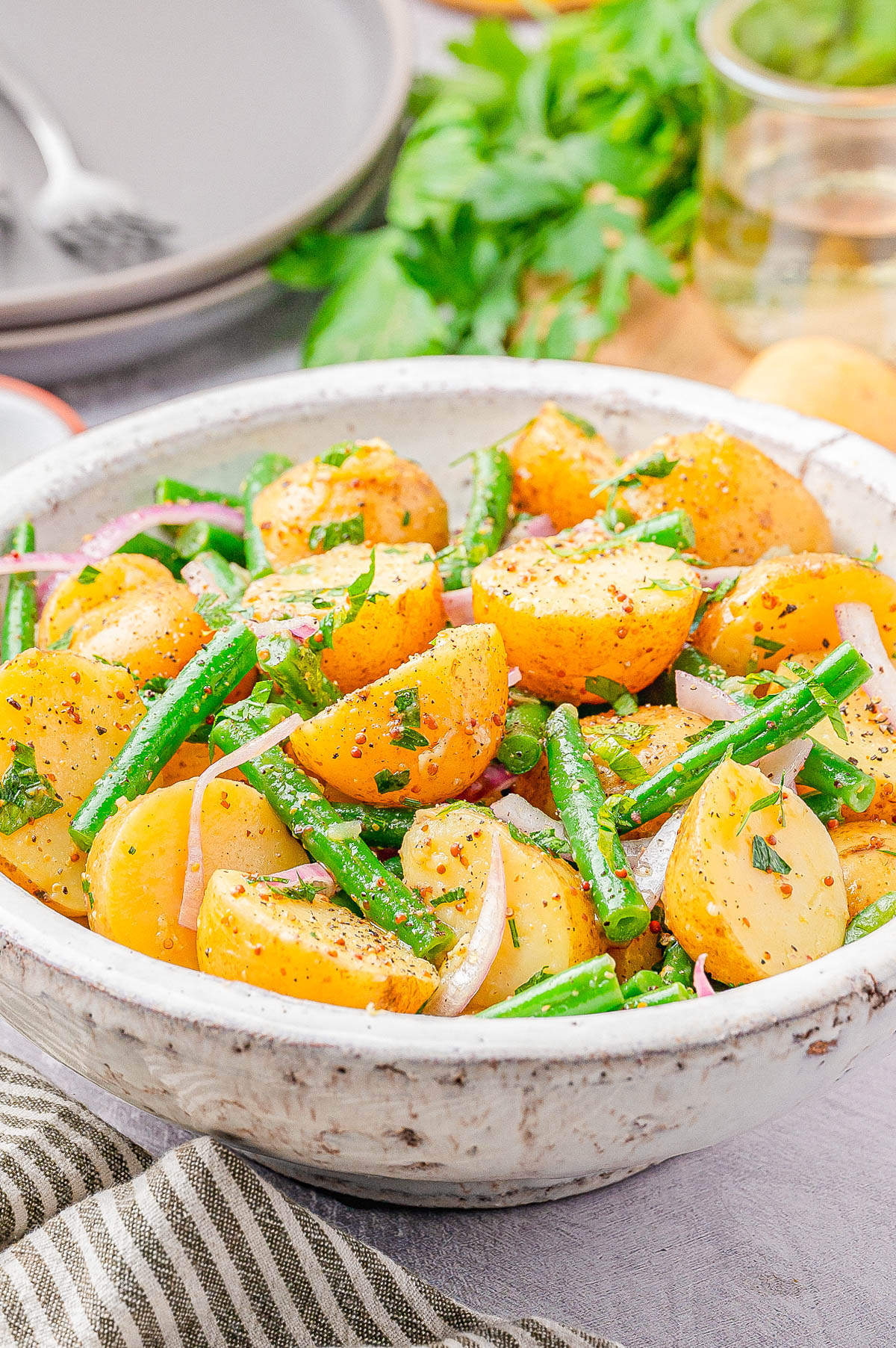 A bowl of potato salad with green beans, sliced red onions, and herbs, seasoned with black pepper, on a table with plates and parsley in the background.