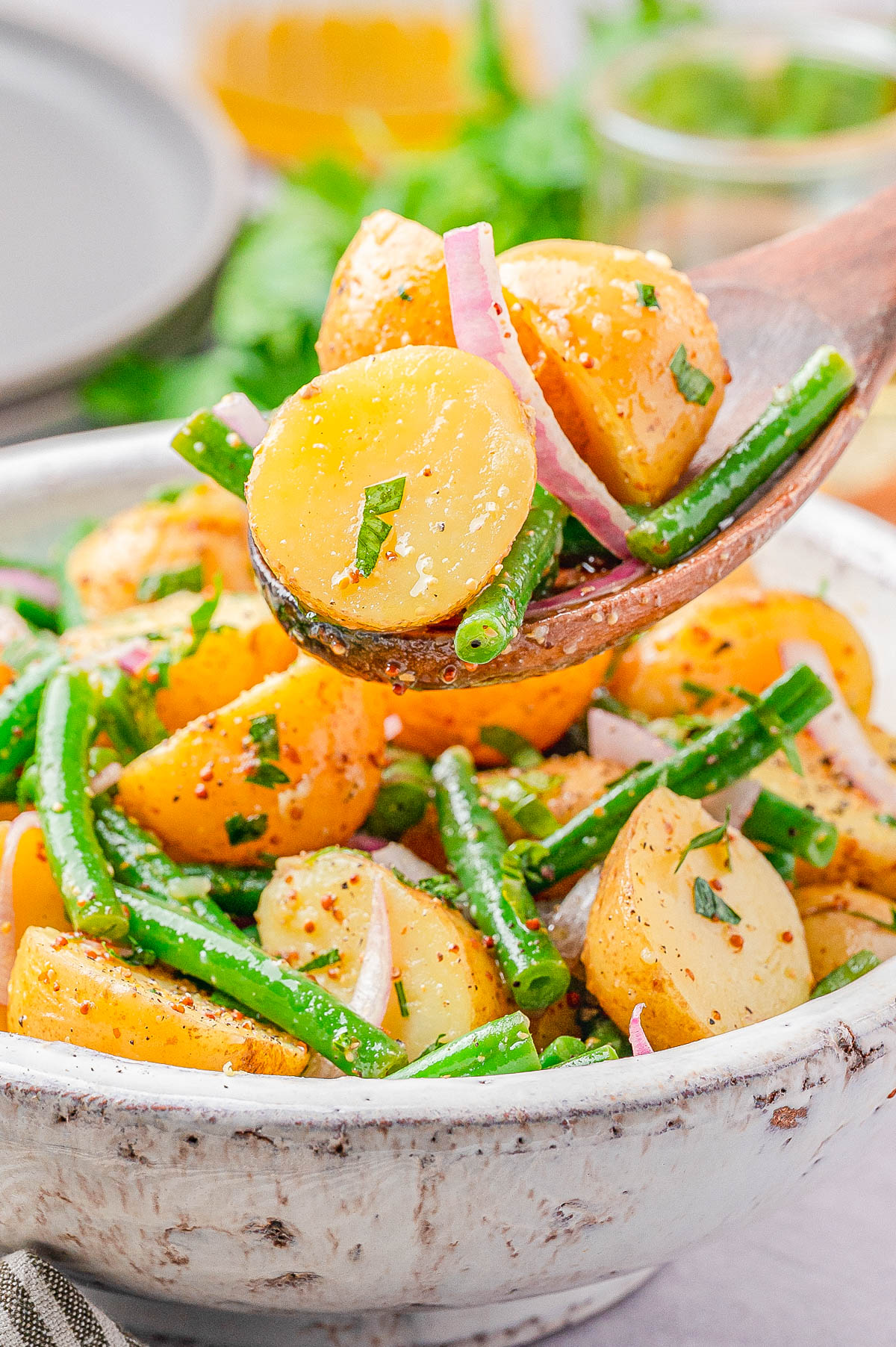 A bowl of potato salad with green beans, red onion slices, and herbs, with a wooden spoon lifting a portion.