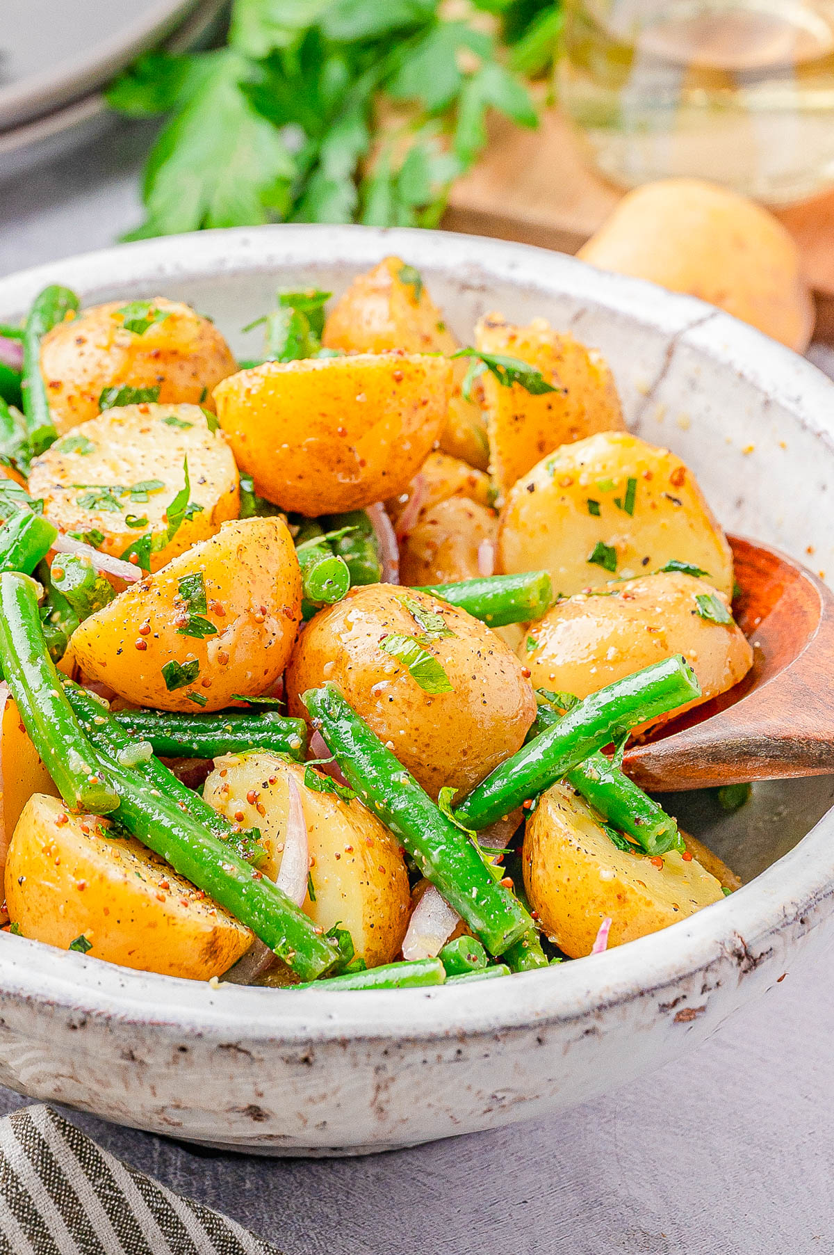 A bowl of potato salad with green beans, red onions, and herbs, served with a wooden spoon.