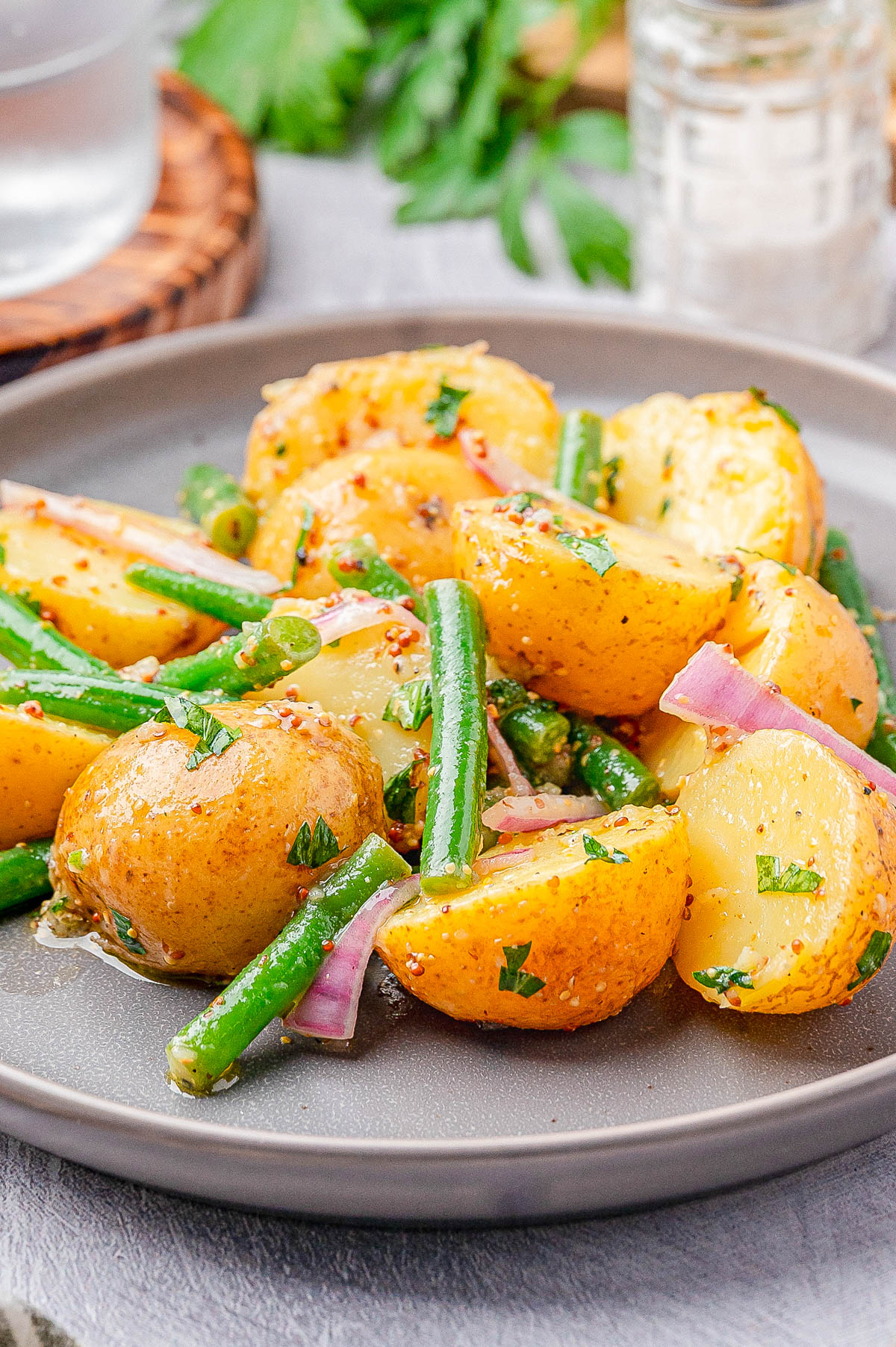 A plate of potato salad with halved baby potatoes, green beans, red onion slices, and chopped parsley, dressed with vinaigrette.