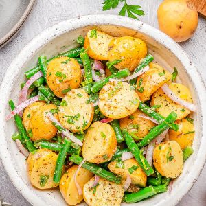 A bowl of potato salad with green beans, red onion slices, and fresh herbs, on a light surface with parsley, a whole potato, and a jar in the background.