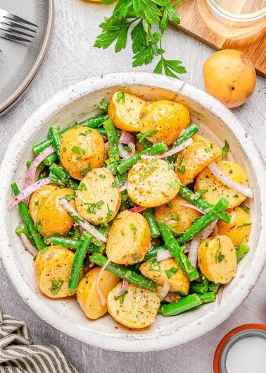 A bowl of potato salad with green beans, red onion slices, and fresh herbs, on a light surface with parsley, a whole potato, and a jar in the background.