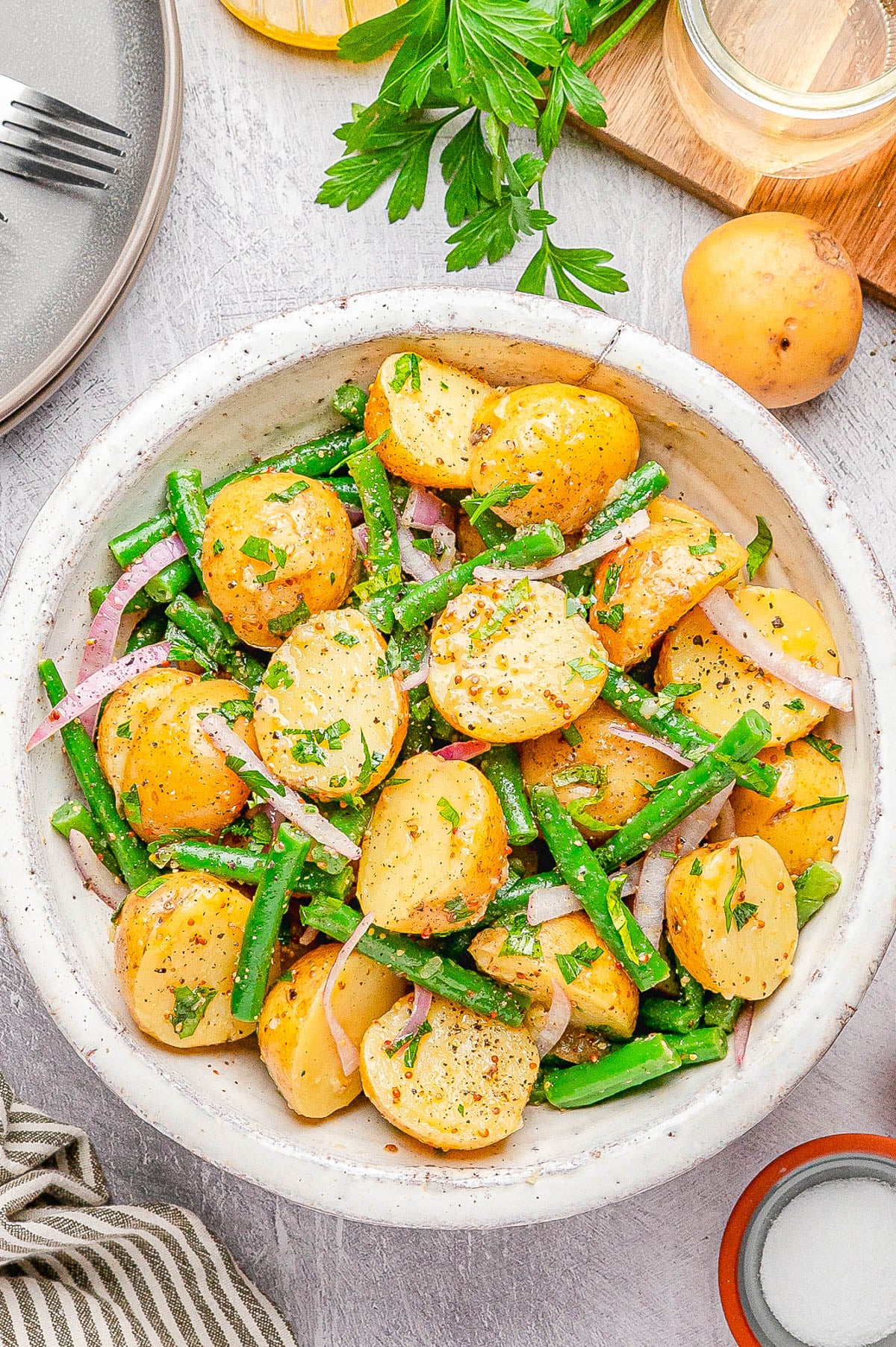 A bowl of potato salad with green beans, red onion slices, and fresh herbs, on a light surface with parsley, a whole potato, and a jar in the background.