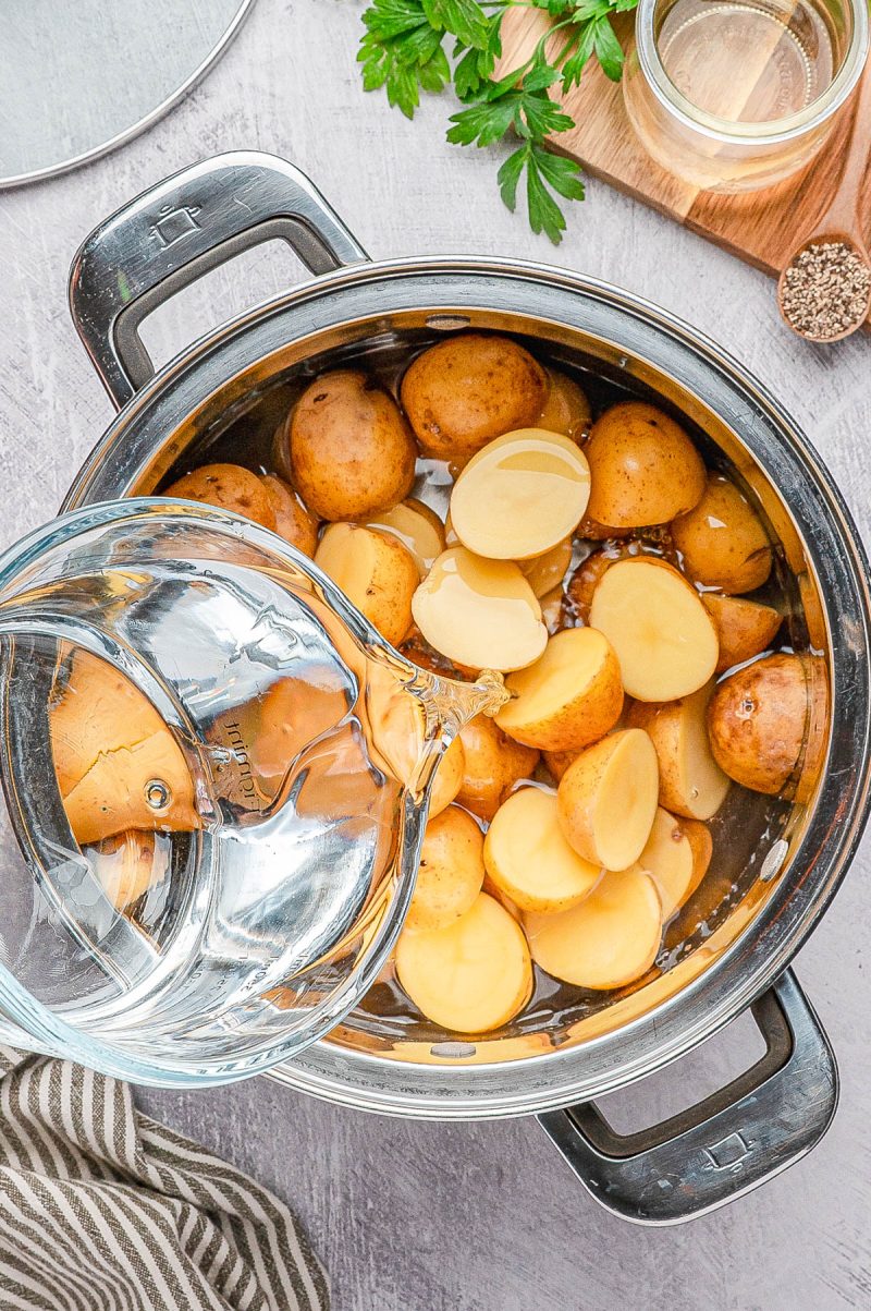 A pot filled with halved and whole potatoes is being topped with water from a glass pitcher on a kitchen countertop.