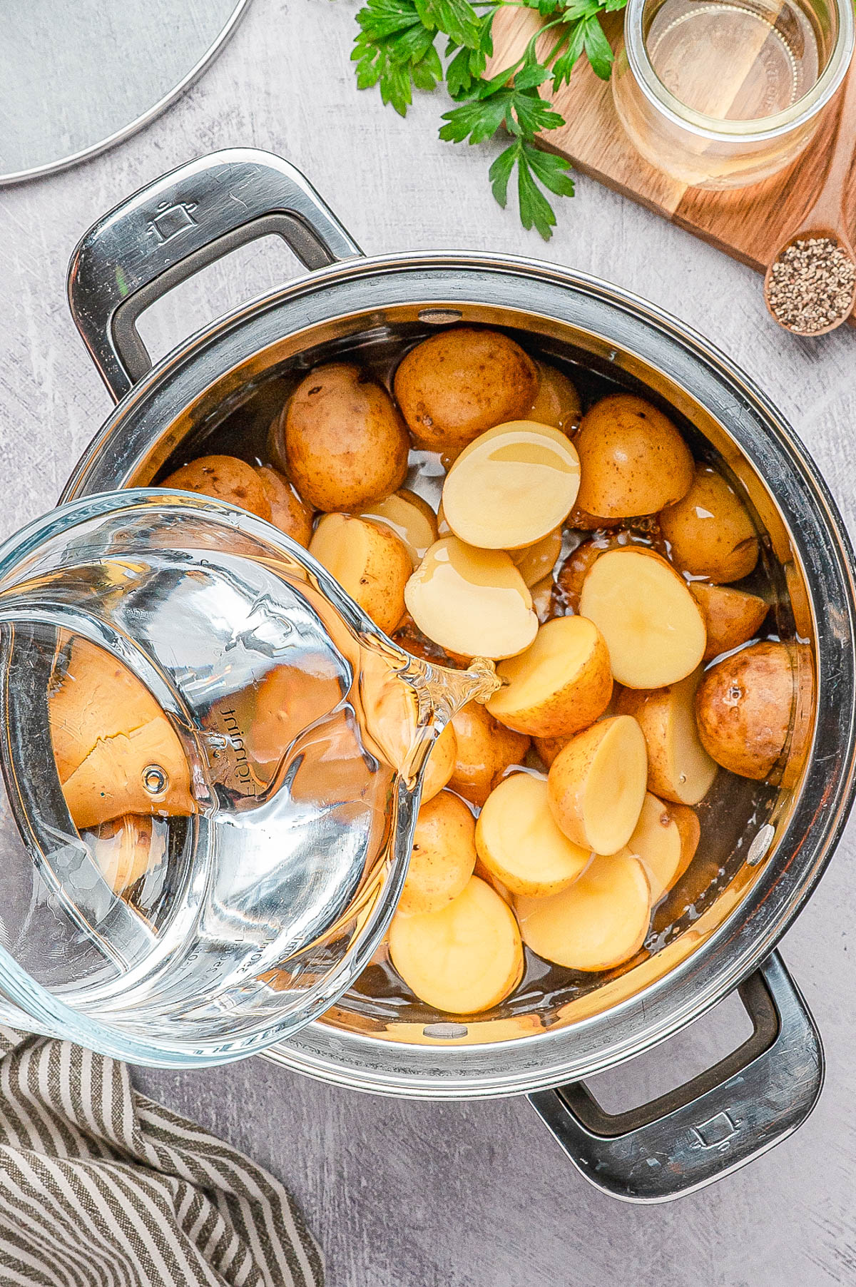 A pot filled with halved and whole potatoes is being topped with water from a glass pitcher on a kitchen countertop.