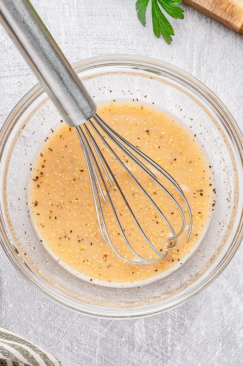 A metal whisk rests in a clear glass bowl containing a light brown, pepper-speckled liquid mixture on a white textured surface.