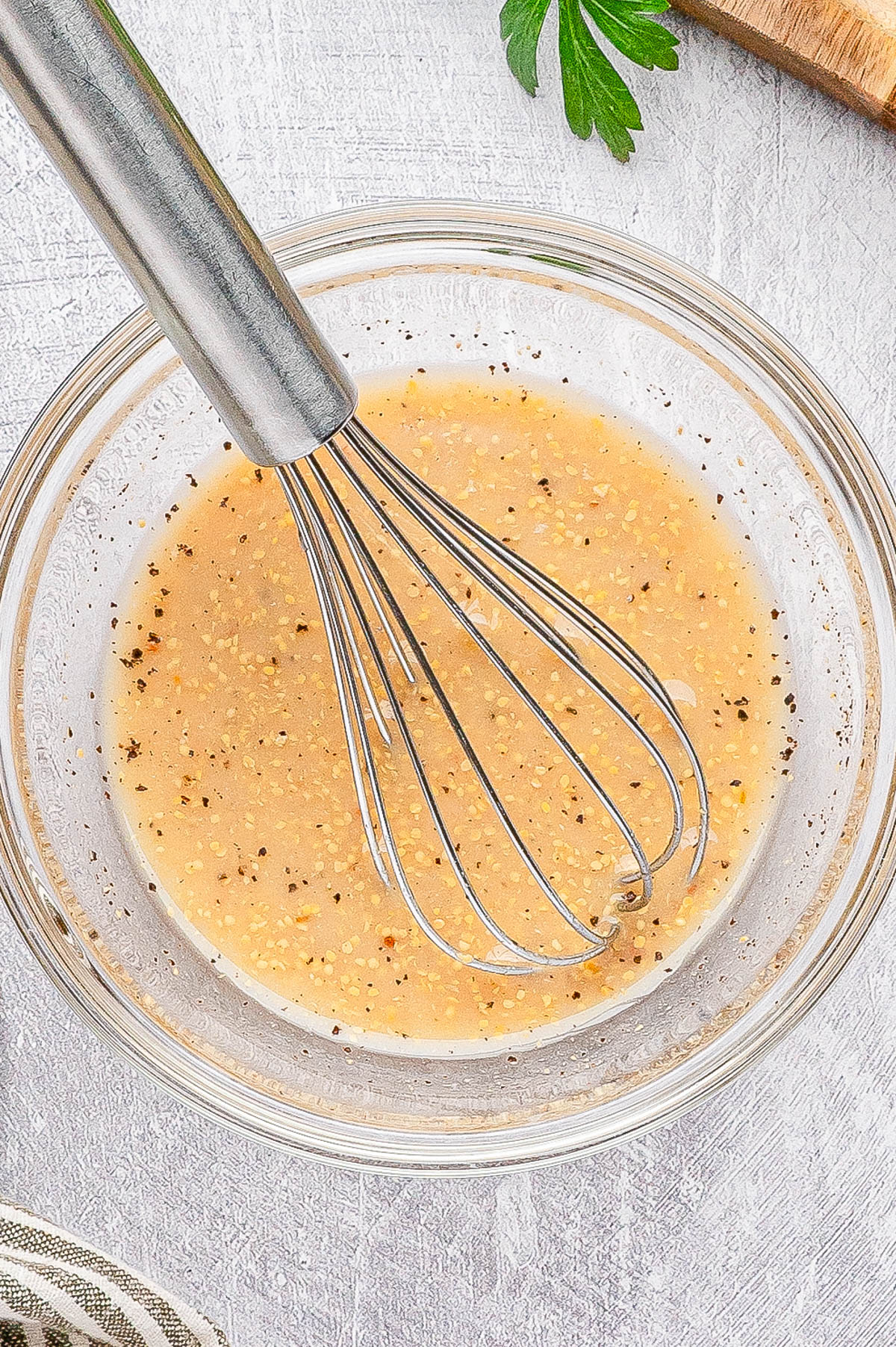A metal whisk rests in a clear glass bowl containing a light brown, pepper-speckled liquid mixture on a white textured surface.