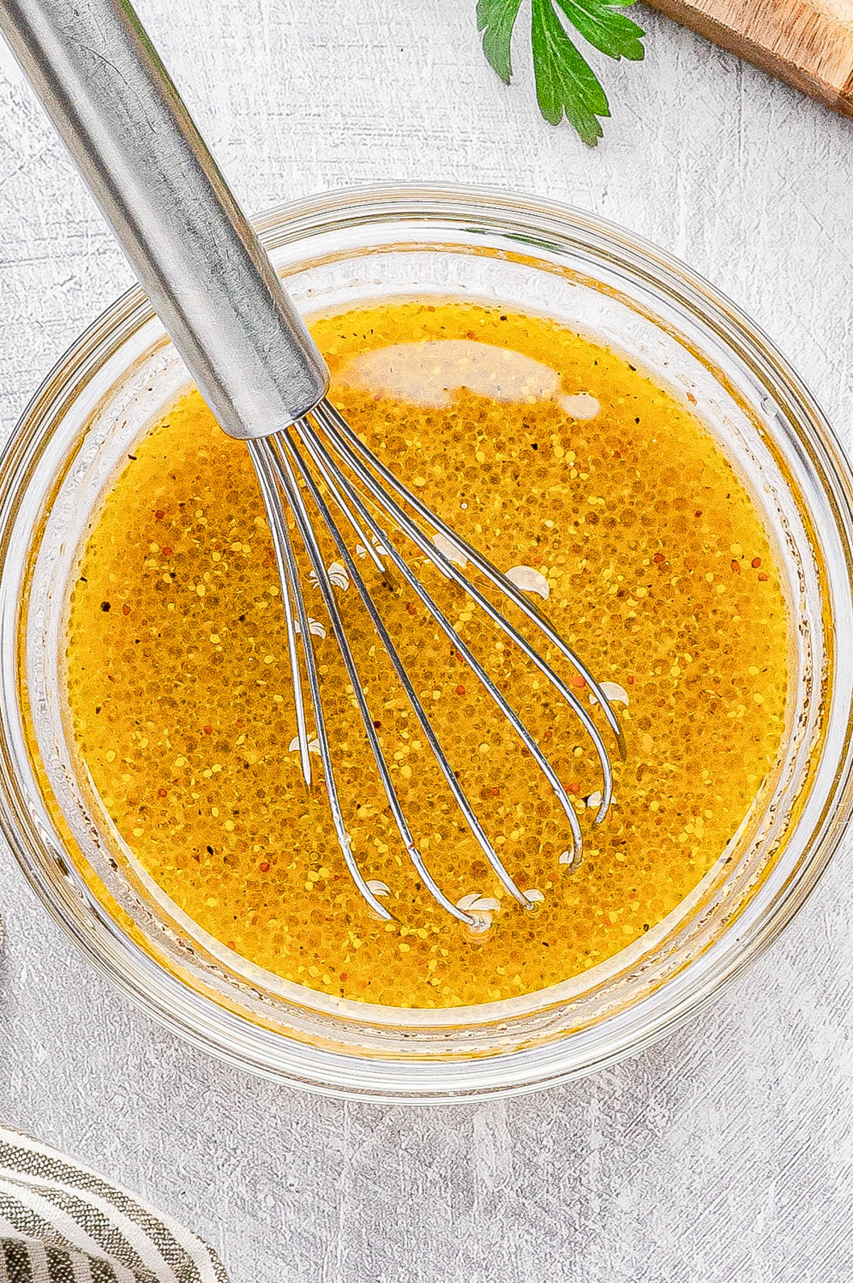 A metal whisk in a glass bowl filled with yellow vinaigrette dressing on a white textured surface.