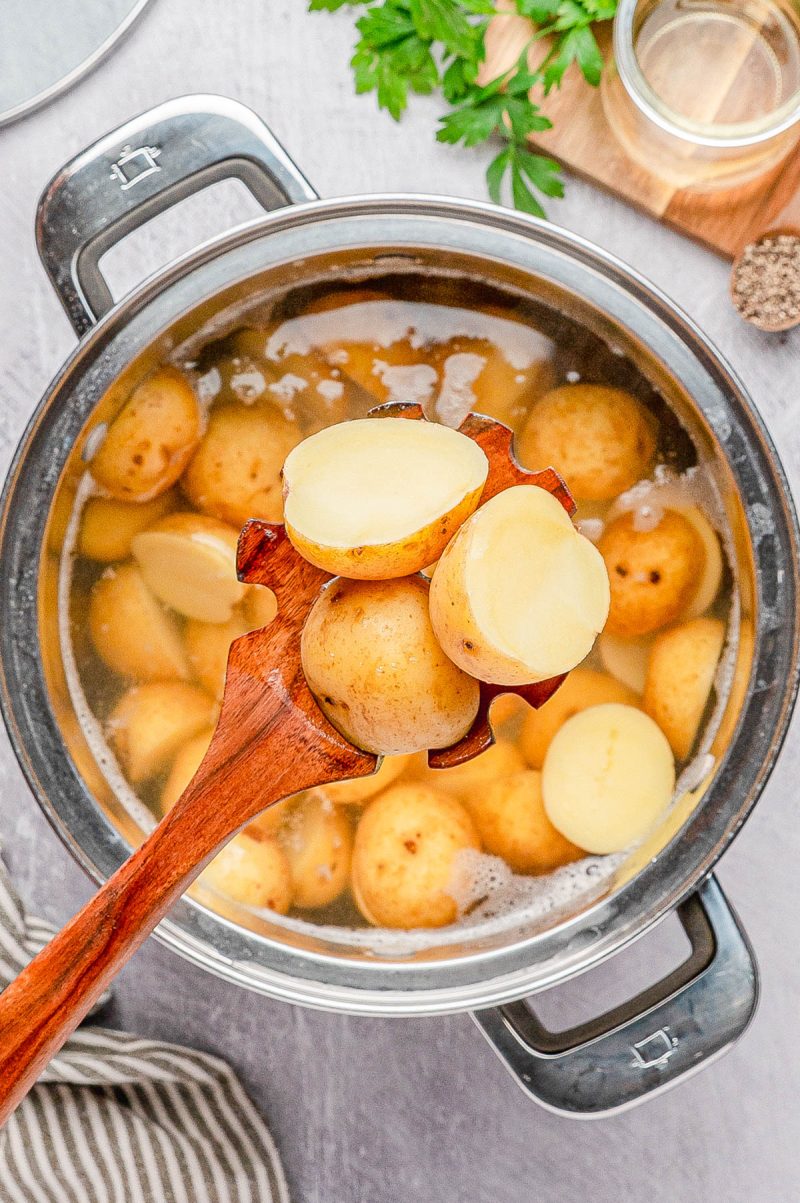 A wooden spoon holds sliced boiled potatoes above a pot filled with whole and halved potatoes in water.