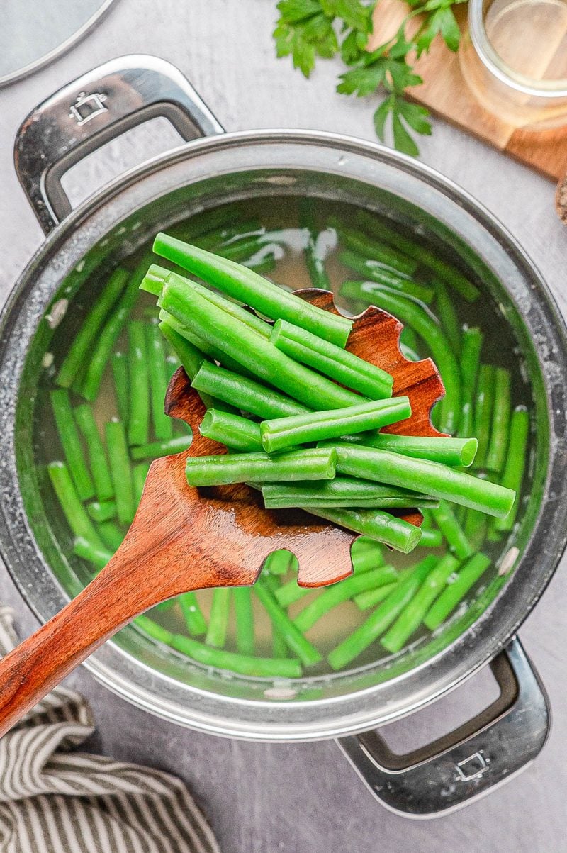 A wooden spoon holds cut green beans above a pot of boiling water with more green beans inside.