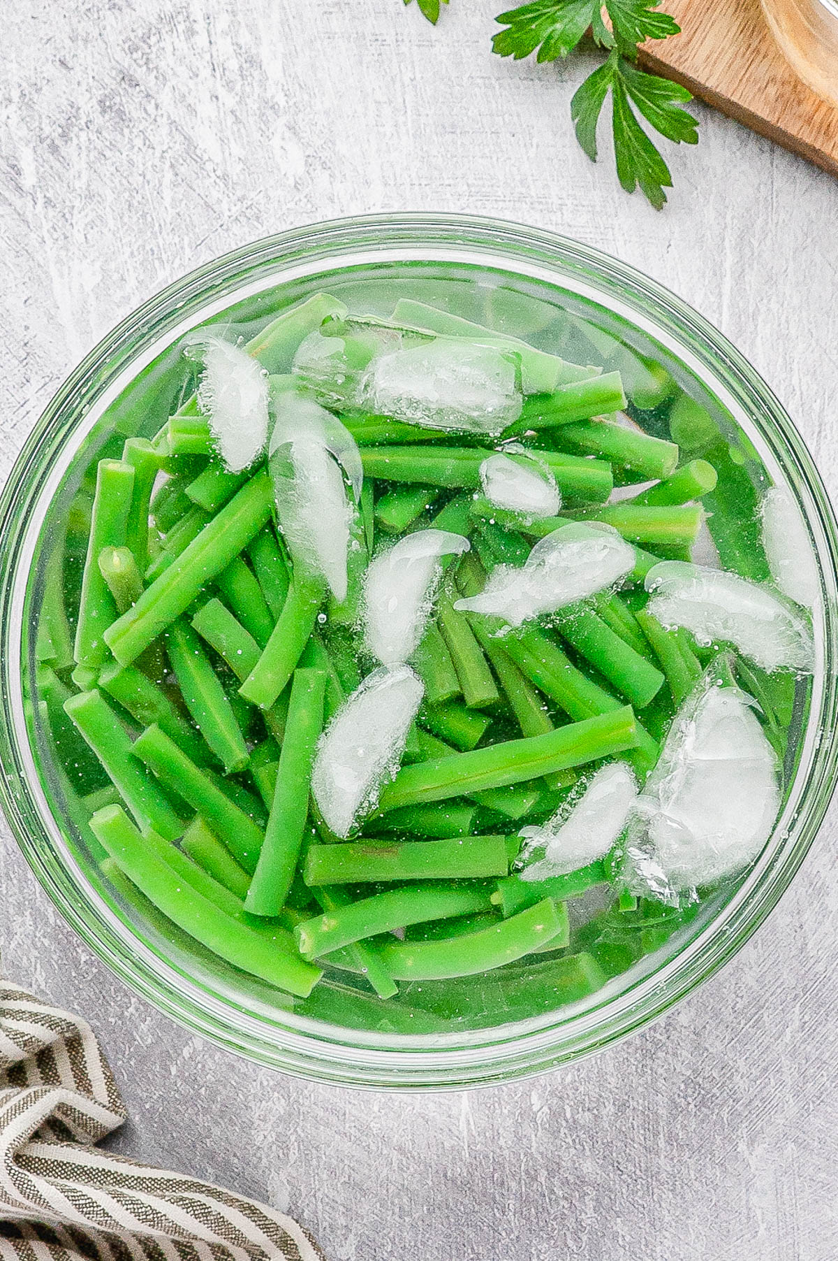 A glass bowl filled with cut green beans and ice cubes submerged in water, placed on a light gray surface.