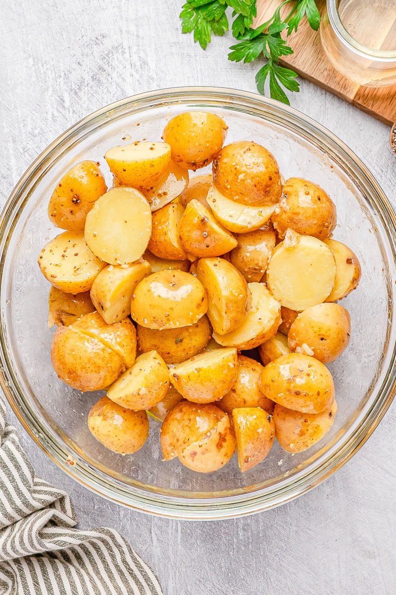 A glass bowl filled with boiled and sliced yellow potatoes, seasoned with herbs, on a light-colored surface with a striped cloth nearby.