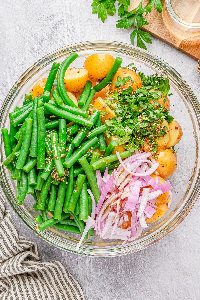 A glass bowl containing halved baby potatoes, green beans, sliced red onions, and chopped herbs on a light surface.