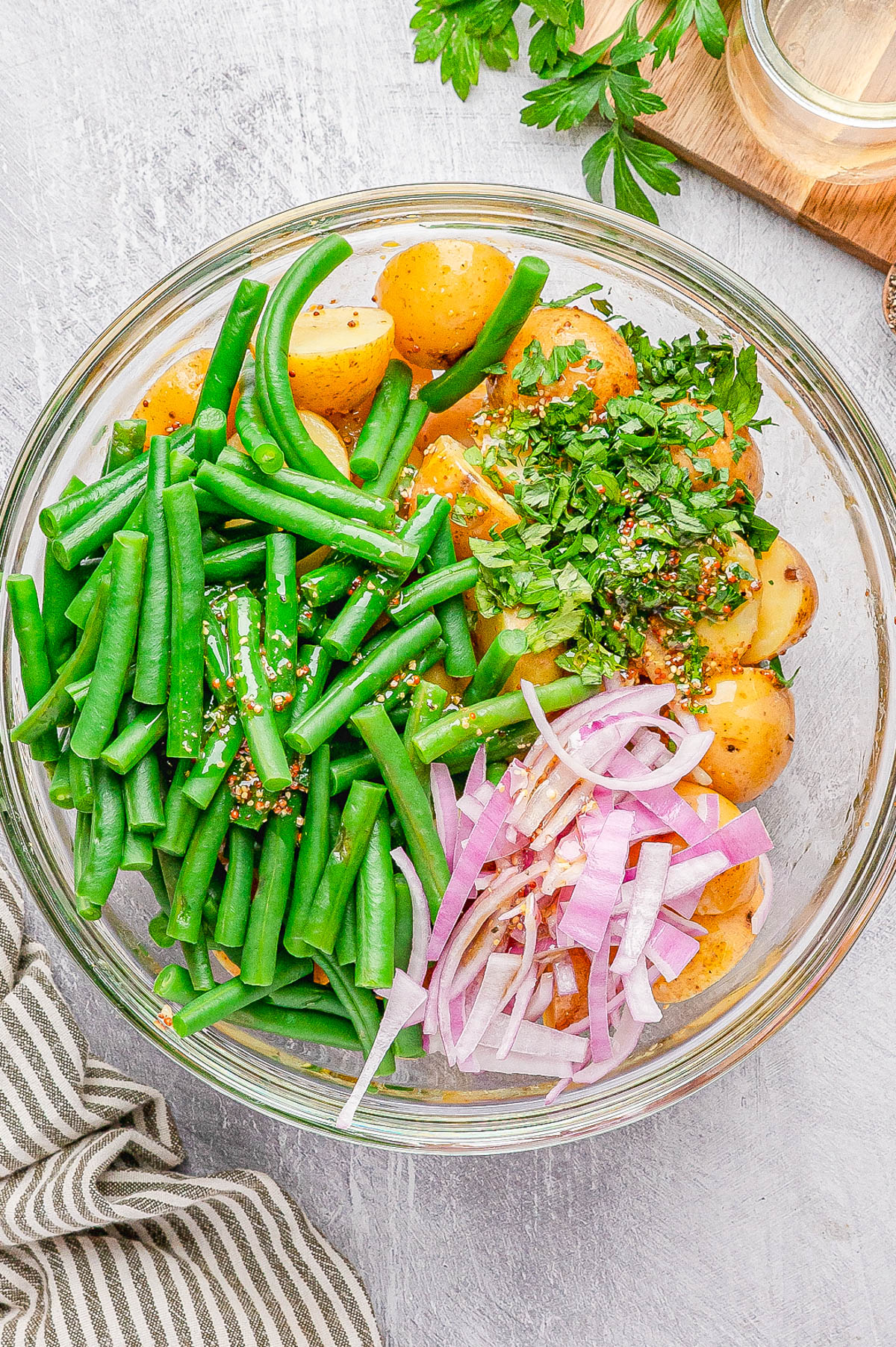 A glass bowl containing halved baby potatoes, green beans, sliced red onions, and chopped herbs on a light surface.