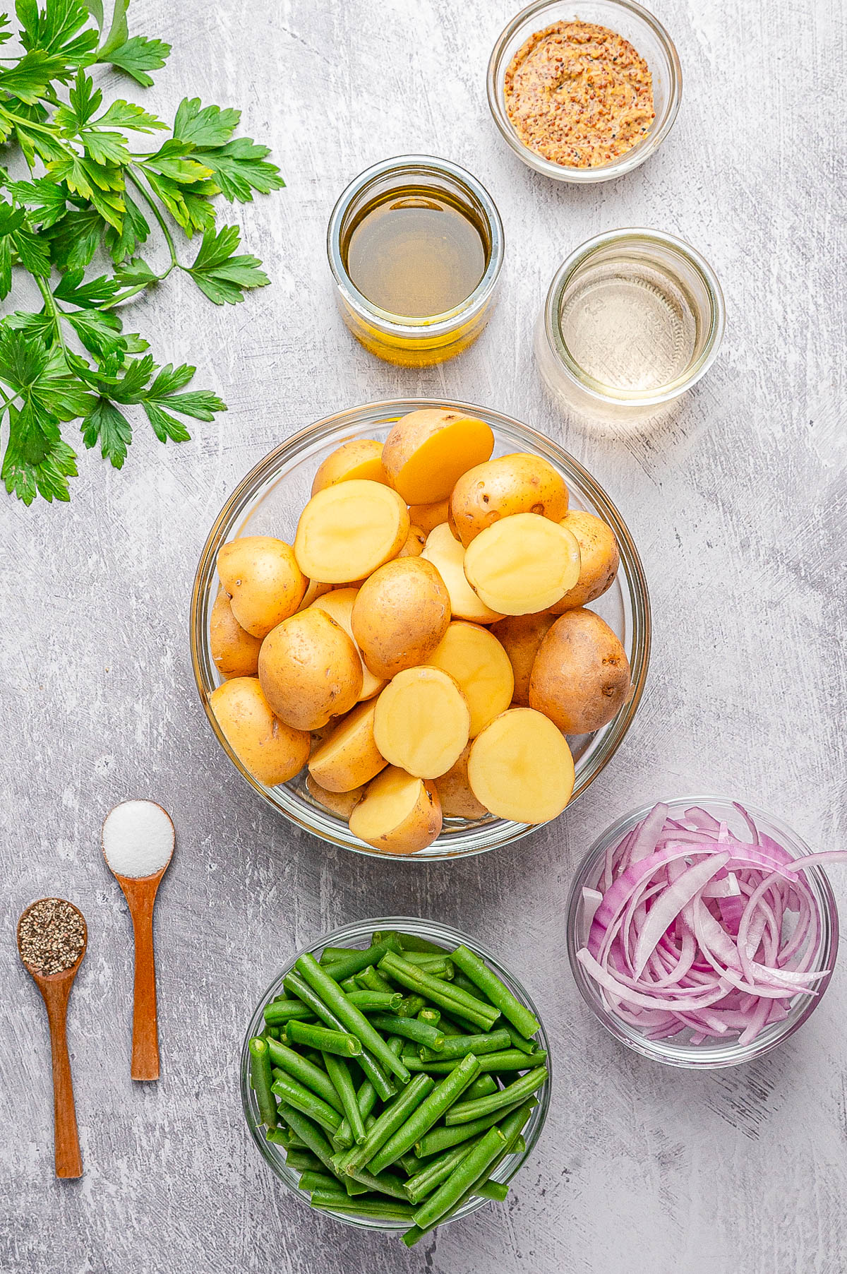 Overhead view of halved potatoes, sliced onions, green beans, parsley, oil, vinegar, mustard, salt, and pepper arranged on a light surface.